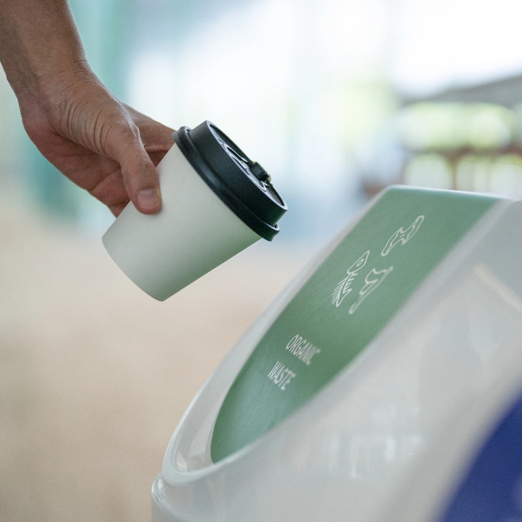 An image of a person throwing a disposable cup into a waste-bin marked with the words 'organic waste'.